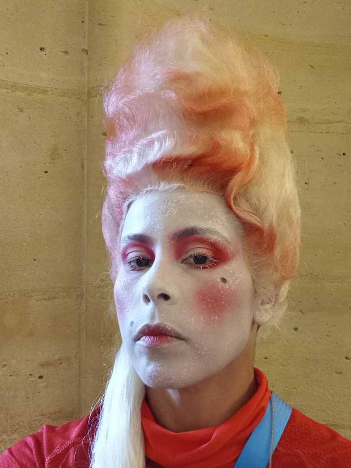 A close-up of Brito's head, showcasing her powdered white makeup and tall white wig with hot pink accents.